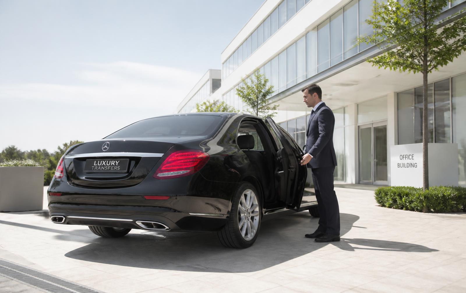 Chauffeur opening the door of a black executive sedan outside a sunlit modern office building